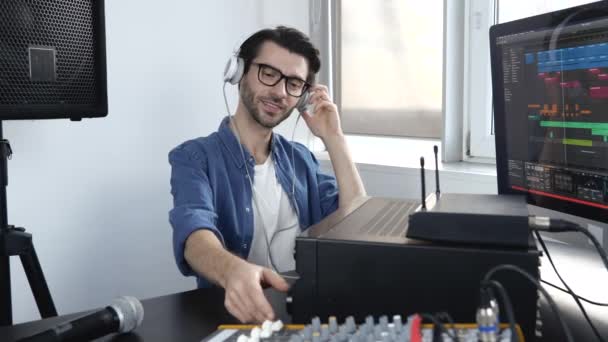 Young man in sound recording room. Guy sit on table and adjusting console in slow motion. Working with professional sound equipment. Listen to music through headphones. - Footage, Video