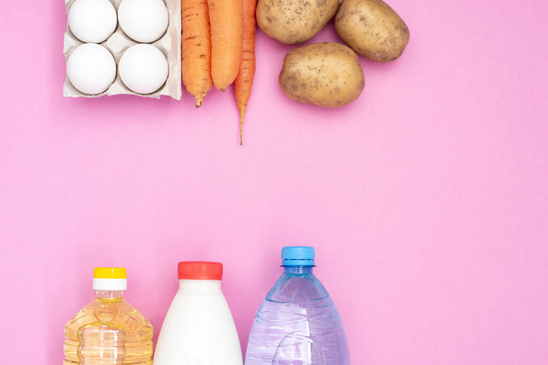 Voedsel donatie voor de behoeftigen op een roze achtergrond bovenaanzicht. Aardappelen, wortelen, water, melk, zonnebloemolie en eieren. - Foto, afbeelding