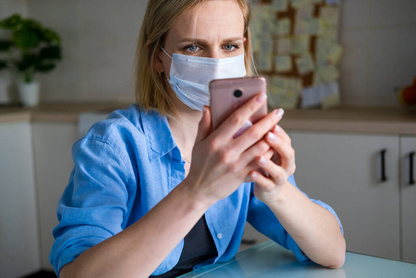 Woman in medical mask is working office work remotely from home on kitchen. Talking mobile phone. Using computer. Distance learning online education and work. - Φωτογραφία, εικόνα