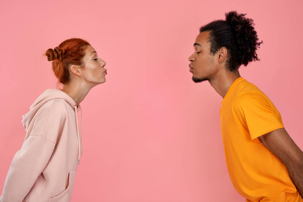 Profile photo of mixed race couple in love face each other, pout lips and going to kiss, close eyes from pleasure, dressed in stylish clothes, have date, over pink background. Romantic relationships - Photo, image