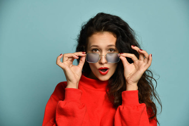 Much surprised adult woman with long curly hair in red sweater looking at us over her stylish rectangular glasses - Photo, image
