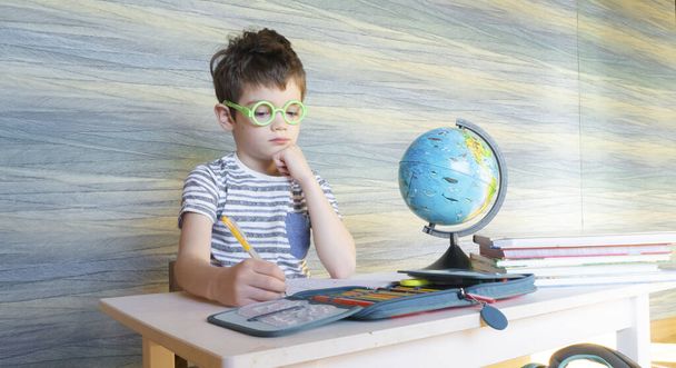 Little egghead schoolboy is doing homework in geography. A primary school student studies in distance home teaching. A student in a school class performs a scheduled test. - Photo, Image