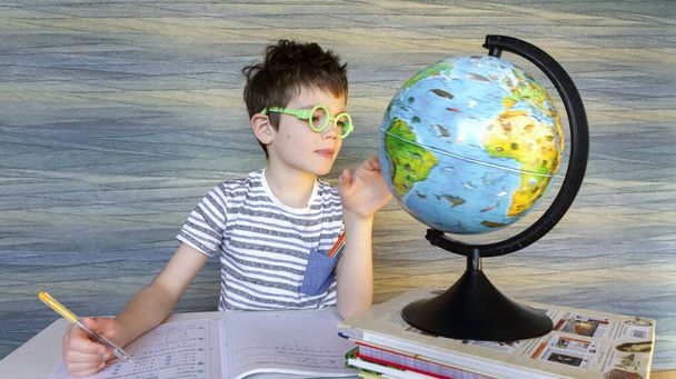 Крутий розумний хлопчик обертає глобус на уроці географії. Молодший студент робить домашнє завдання, дивлячись на земну кулю з інтересом. Триває навчання на домашній дистанції
. - Фото, зображення