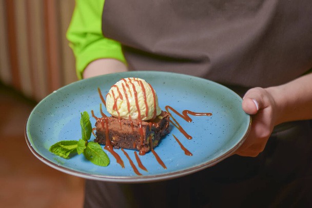 Ober serveert chocoladebrownie met vanille-ijs en chocoladekaramelsaus in een blauw bord. Heerlijk dessert. - Foto, afbeelding