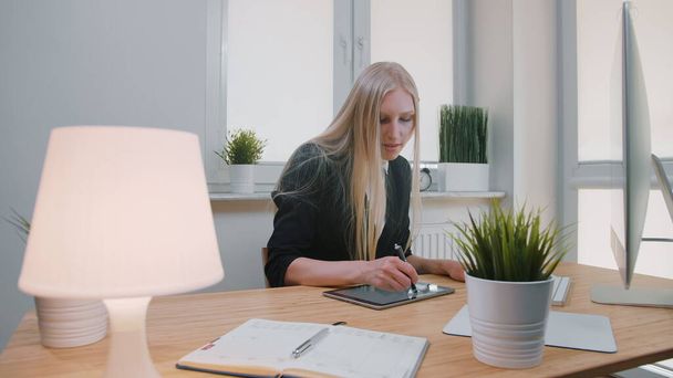 İş kadınları ofiste tablet üzerinde çalışıyor. Zarif takım elbiseli, uzun saçlı çekici sarışın kadın ahşap masada bilgisayarla oturuyor ve ışıklı ofisteki kalemle tablet üzerinde çalışıyor.. - Fotoğraf, Görsel