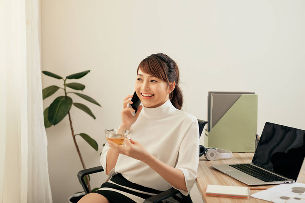 Jovem mulher asiática falando ao telefone enquanto bebe chá e está sentada no escritório
 - Foto, Imagem