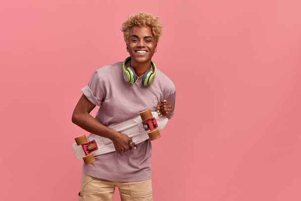 Happy black woman with blonde hairdo holds skateboard, has fun with friend while skating, enjoys active rest, wears lilac t shirt and wide pants, poses over pink background. People, hobby, leisure - Valokuva, kuva