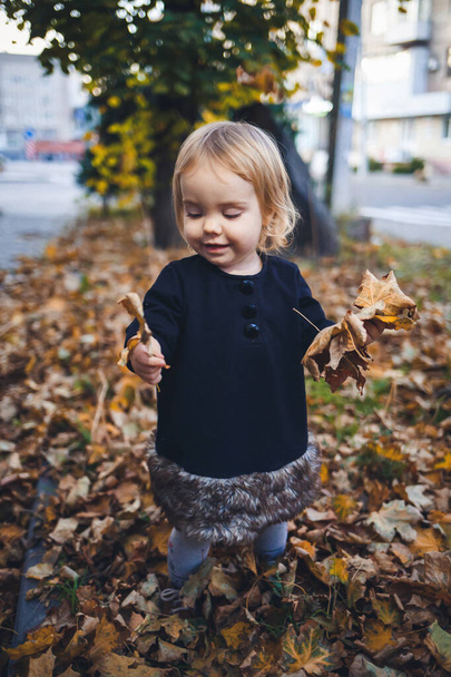 Una niña de 1.5 años está de pie y sonriendo. El niño lanza hojas. Chica alegre en el bosque de otoño
 - Foto, imagen