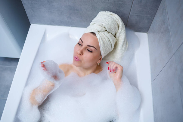 Girl woman with a white towel on her head lies in the bathroom in a white bath. In the hands of a lot of muddy soap. Full whirlpool foam. Relax after a hard day. Spa relaxing treatment - Photo, Image