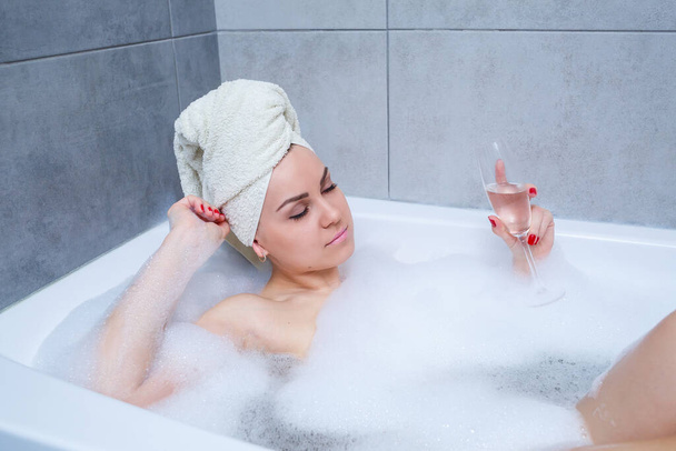 Девушка с белым полотенцем на голове лежит с бокалом шампанского в белой ванне. В ее руках много мыла для мочи. Полная пена от водоворота. Расслабься после тяжелого дня. СПА процедуры
 - Фото, изображение