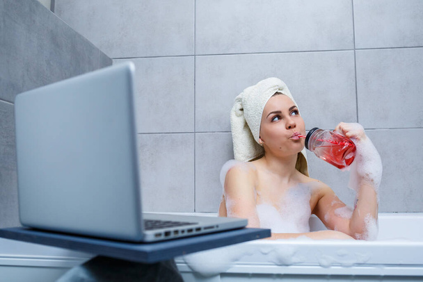Eine junge Frau mit einem Handtuch auf dem Kopf trinkt Saft und schaut sich einen Film auf einem Laptop an, während sie in einem Bad in einem Schönheitssalon sitzt. Entspannen Sie im Badezimmer ohne Bettwäsche. Körperpflege und Entspannungskonzept. - Foto, Bild