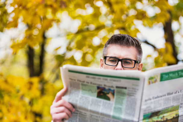 Portrait of mature attractive man sitting on bench while reading newspaper in park - Foto, Imagem
