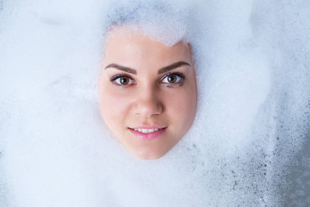Szoros portré egy lányról fürdőkádban és fehér hab az arca körül. Különböző érzelmek, grimaszok, női mosoly. - Fotó, kép