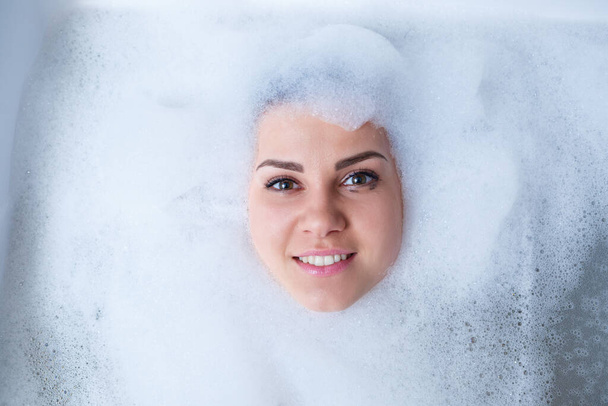 Closeup portrait of a girl in a bathtub and white foam around her face. Different emotions, grimaces, woman smile - Fotografie, Obrázek