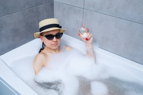 Beautiful young woman in a hat on her head and sunglasses drinking a cocktail takes a bath at home. Relax after a hard day. The spa is a relaxing procedure. - Photo, Image