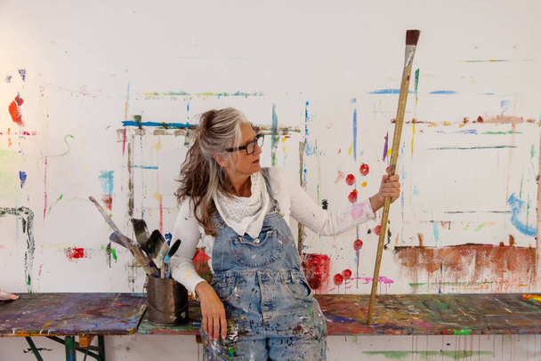 proud older artist woman, in her fifties with grey hair and black glasses holds a very big paintbrush - 写真・画像