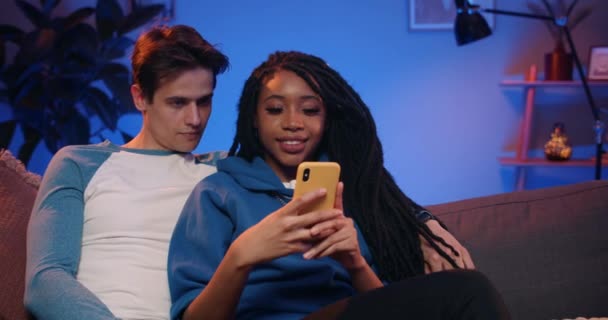 Handsome young man scrolling news feed while sitting on sofa near his girlfriend at living room. Millennial cheerful couple using smartphone while spending time at home. Concept of leisure. - Video