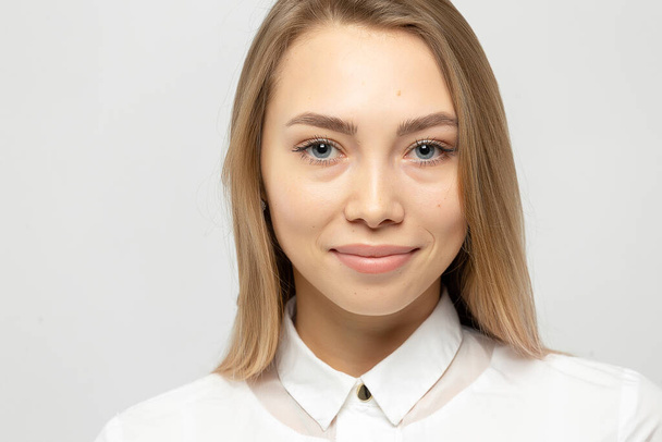 Close-up portrait of beautiful happy young attractive blonde woman in a white shirt with clean skin looking at camera against gray-white background - Photo, Image