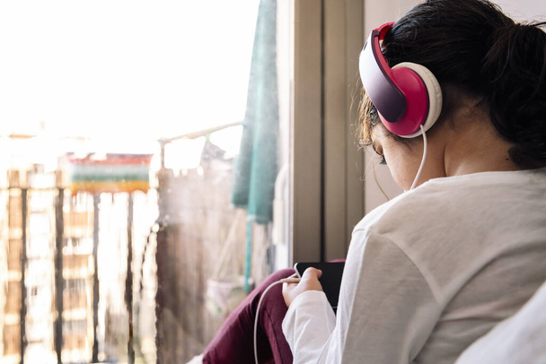 little girl sitting in front of her house balcony with headset watching videos on the phone, home entertainment for children concept, copy space for text - 写真・画像