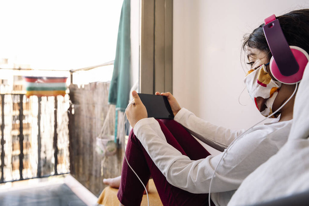 little girl sitting in front of her home window with a headset and protective mask watching videos on the phone, coronavirus concept and home entertainment for children, copy space for text - Foto, Imagem