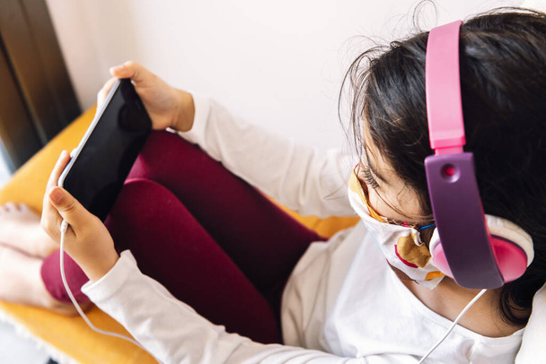 little girl sitting at home with a headset and protective mask learning with the phone, coronavirus concept and home entertainment for children - Valokuva, kuva