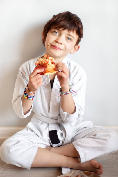 Retrato vertical de la linda chica de 7 años hambrienta de pelo corto en kimono blanco comiendo pizza después de un duro entrenamiento de taekwondo de karate en línea. El niño después del entrenamiento a distancia come en casa. Artes marciales
. - Foto, Imagen