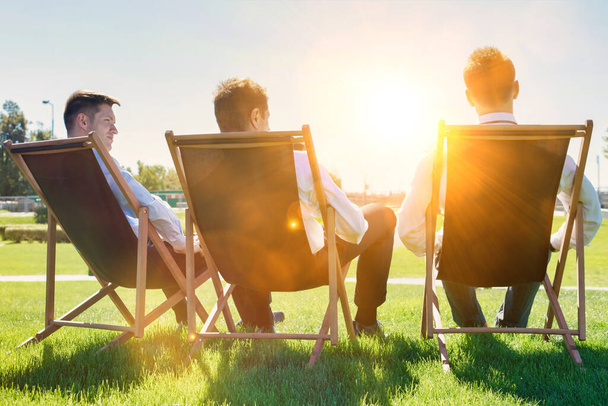 Group of handsome businessmen relaxing and talking about life while sitting on folding chairs at park - Fotoğraf, Görsel