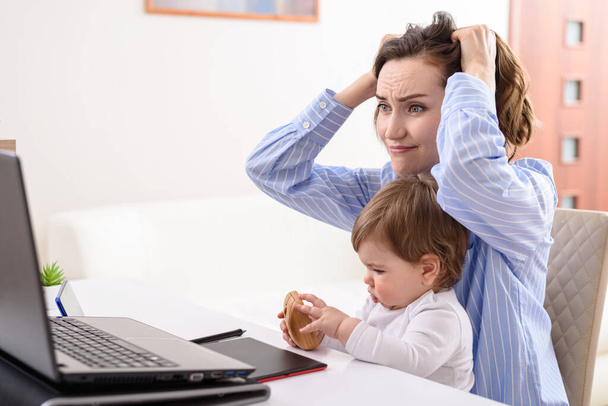 mulher morena em camisa azul com criança no colo trabalhando remotamente no laptop em licença maternidade em casa, falta de tempo, conceito de prazos apertados
 - Foto, Imagem