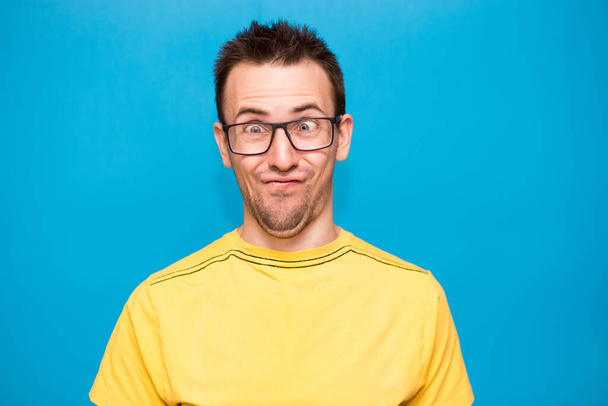 Funny comic man in glasses makes grimace with crosses eyes. Young man in yellow t-shirt with crazy expression has fun alone, plays fool isolated over blue background - 写真・画像