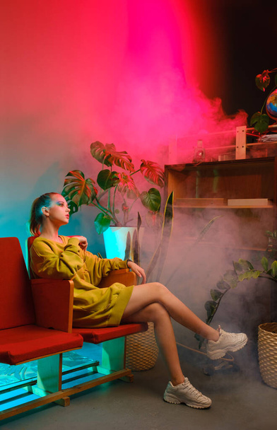A young attractive girl with bright makeup and in colored clothes sits on a red armchair against a fashionable neon background. - Photo, Image