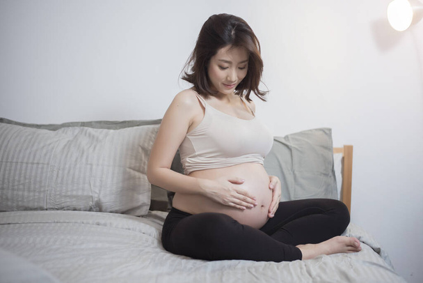 Beautiful asian pregnant woman sit on bed and touching her belly  - Fotoğraf, Görsel