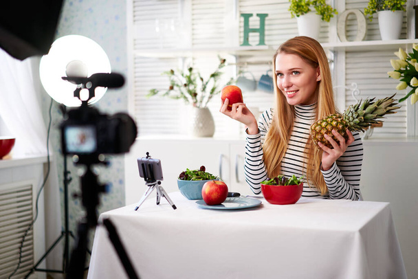 Food blogger cooking fresh vegan salad of fruits in kitchen studio, filming tutorial on camera for video channel. Female influencer holds apple, pineapple and talks about healthy eating. Fructorianism - Photo, image