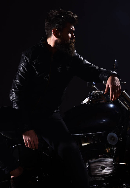 Man with beard, biker in leather jacket lean on motor bike in darkness, black background. Macho, brutal biker in leather jacket stand near motorcycle at night time. Brutality and masculine concept. - Photo, image