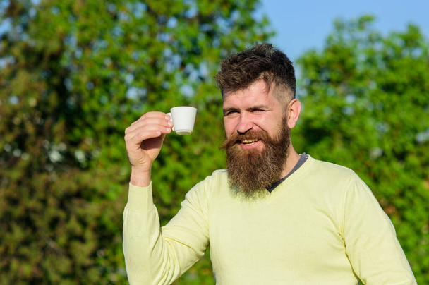 Man with long beard enjoy coffee. Man with beard and mustache on smiling face drinks coffee at balcony, tree on background, defocused. Morning ritual concept. Bearded man with mug drinks coffee. - Foto, afbeelding