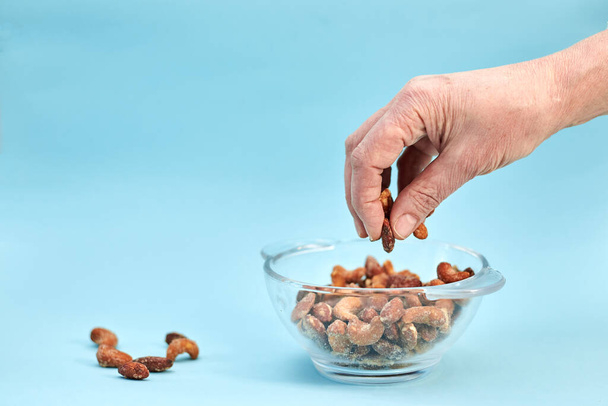 A hand is taking a little bit of nuts from the glass bowl, couple of nuts are laying aside, on light blue background - Photo, Image