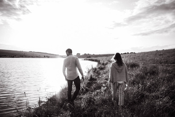 Krásný milující pár kráčející po břehu, objímající se, usmívající se na pozadí oblohy v poli. Ten chlap a ta holka hipsterka cestují - Fotografie, Obrázek