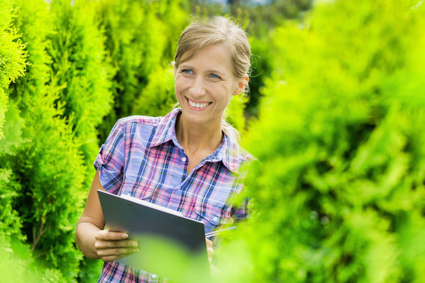 Portrait of mature owner smiling while writing report on clipboard in garden - Photo, Image