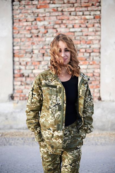 Young curly blond military woman, wearing ukrainian military uniform, posing for picture. Three-quarter portrait of female army soldier standing in front of brick wall of ruined building, smiling. - Photo, Image