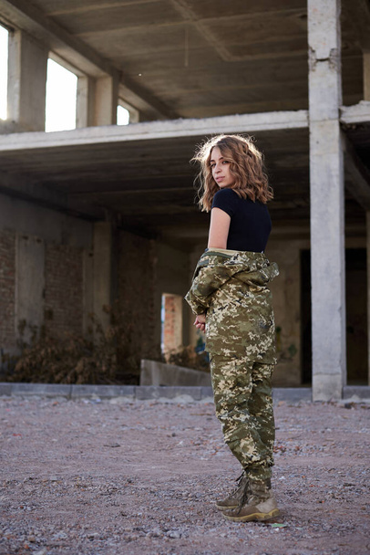 Young curly blond military woman, wearing ukrainian army military uniform and black t-shirt. Full-length portrait of female soldier walking in rim light glare in front of ruined abandoned building. - Fotoğraf, Görsel