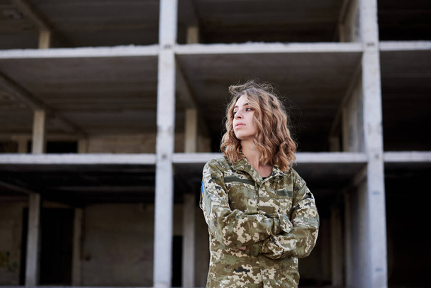 Young curly blond military woman, wearing ukrainian army military uniform and black t-shirt, posing. Three-quarter portrait of female soldier in front of ruined abandoned building,construction site. - Fotoğraf, Görsel