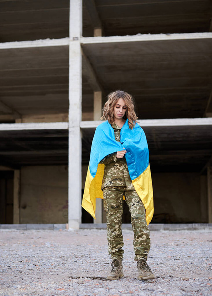 Mladá kudrnatá blondýnka v uniformě ukrajinské armády, pokrytá ukrajinskou modrou a žlutou vlajkou. Plný portrét vojačky před zničenou opuštěnou budovou. - Fotografie, Obrázek