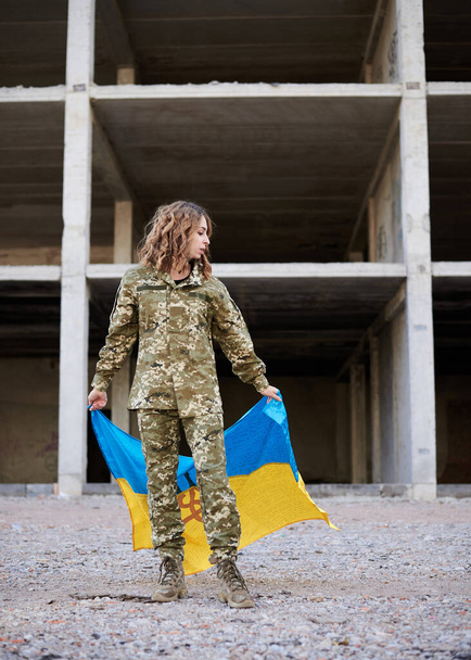 Young curly blond military woman, wearing ukrainian army uniform, holding ukrainian blue and yellow flag. Full-length portrait of female soldier in front of ruined abandoned building. - Foto, afbeelding