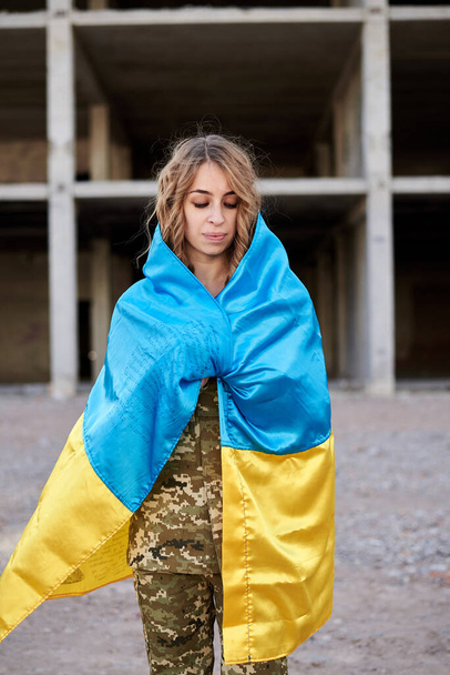 Young curly blond military woman, wearing ukrainian army uniform, covered with ukrainian blue and yellow flag. Three-quarter portrait of female soldier in front of ruined abandoned building. - Zdjęcie, obraz