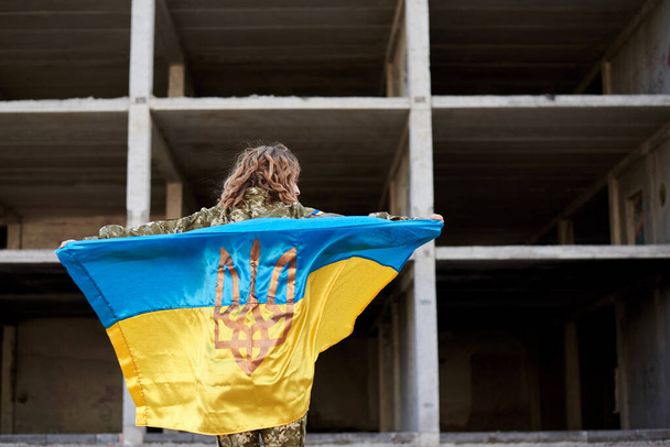 Young curly blond military woman, wearing ukrainian army uniform, holding ukrainian blue and yellow flag. Three-quarter picture from back of female soldier in front of ruined abandoned building. - 写真・画像