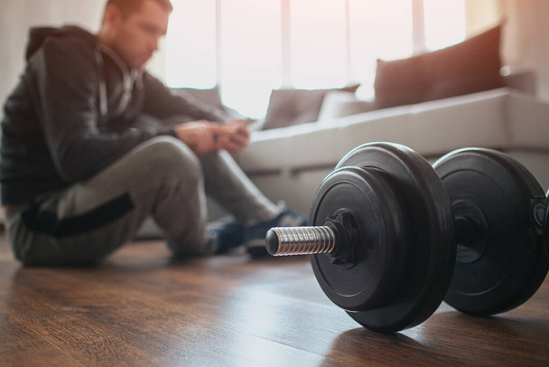 Young ordinary man go in for sport at home. Cut view with blurred background where workout beginner sit on floor and has rest after hard workout. Big dumbbells on floor. Sport amateur. - Φωτογραφία, εικόνα