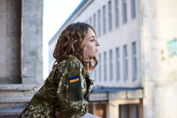 Young curly blond military woman,wearing ukrainian army uniform and black t-shirt. Three-quarter portrait of female soldier inside abandoned building, leaning on windowsill, thinking, looking away - Foto, Imagem