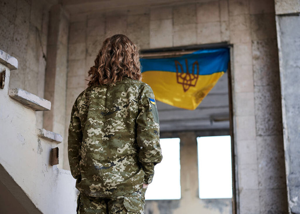 Young curly blond military woman,wearing ukrainian army uniform and black t-shirt. Three-quarter back portrait of female soldier inside ruined building with ukrainian blue and yellow flag at the back. - Foto, afbeelding