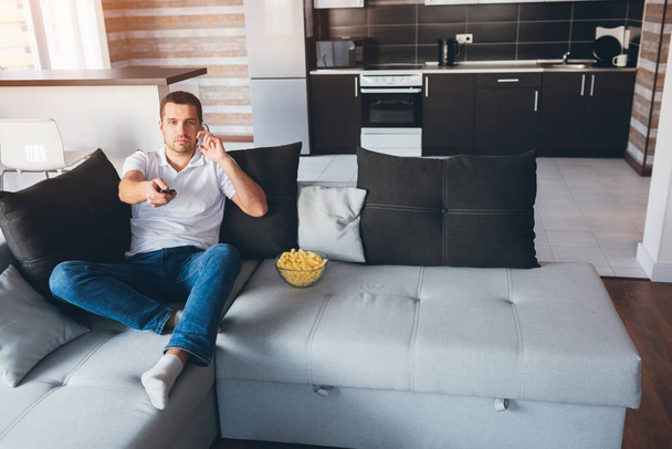Young man watch tv in his own apartment. Busy guy sitting on couch and talking on phone. Switching tv channels with remote control. Entertain himself. Eat snacks and have fun. - Photo, Image
