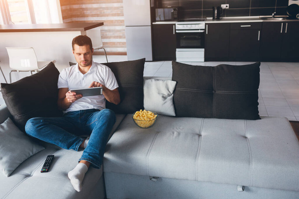 Young man watch tv in his own apartment. Sit alone on couch and work on tablet. Touch screen. Remote work at home. Alone in room concentrated hardworking person. Daylight. - Фото, зображення