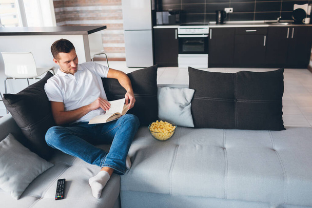 Young man watch tv in his own apartment. Picture of handsome smart guy reading book in room alone. Studying or reading for peasure. After watching movie. Relax and rest. - Photo, Image